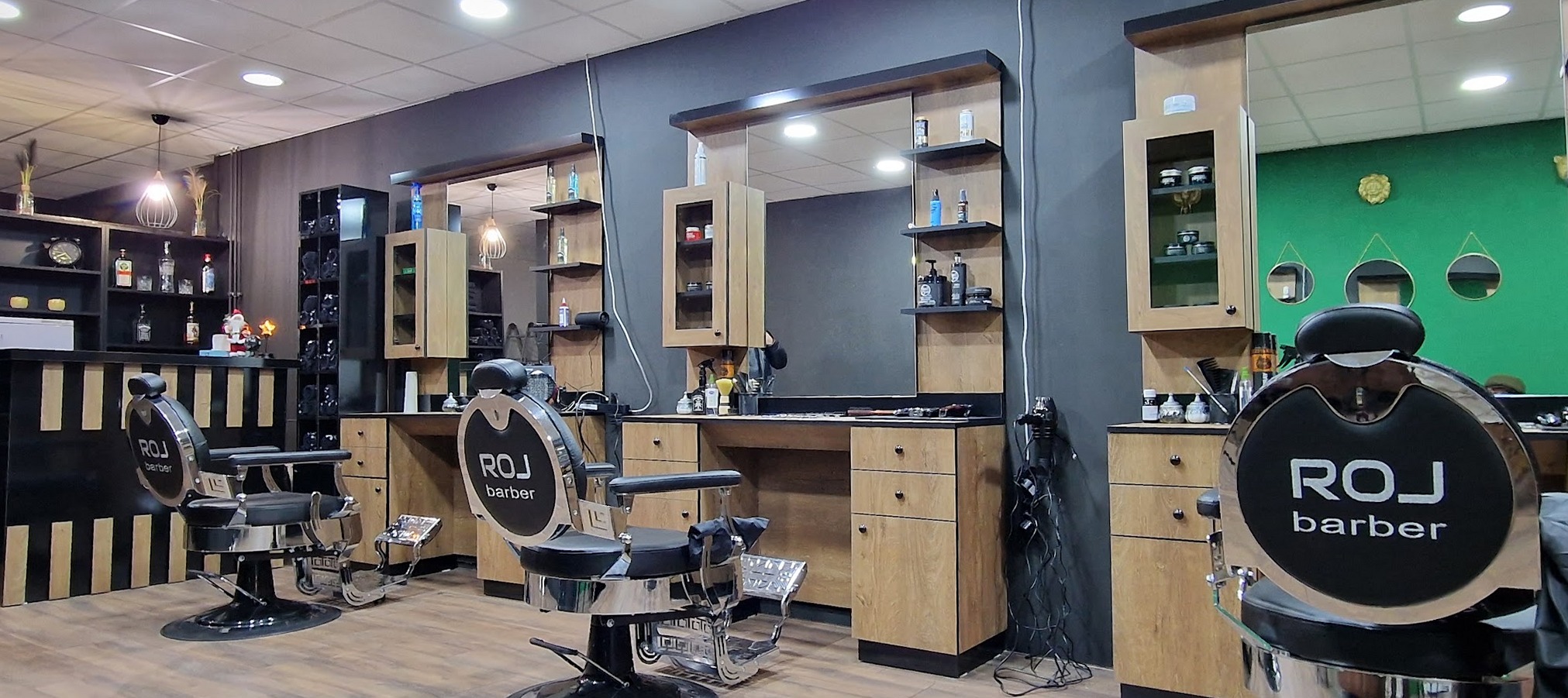 De zaak van Roj Barber: drie werkplekken met eikenhouten kasten en spiegels, zwarte stoelen met het ROJ barber logo op de rugleuning en de groene muur